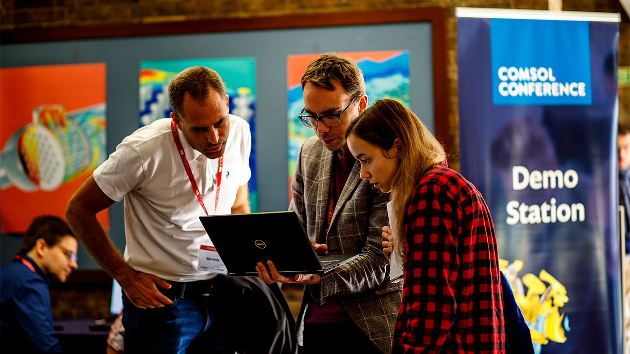 Three conference attendees looking at a laptop together in front of a COMSOL Conference demo station.
